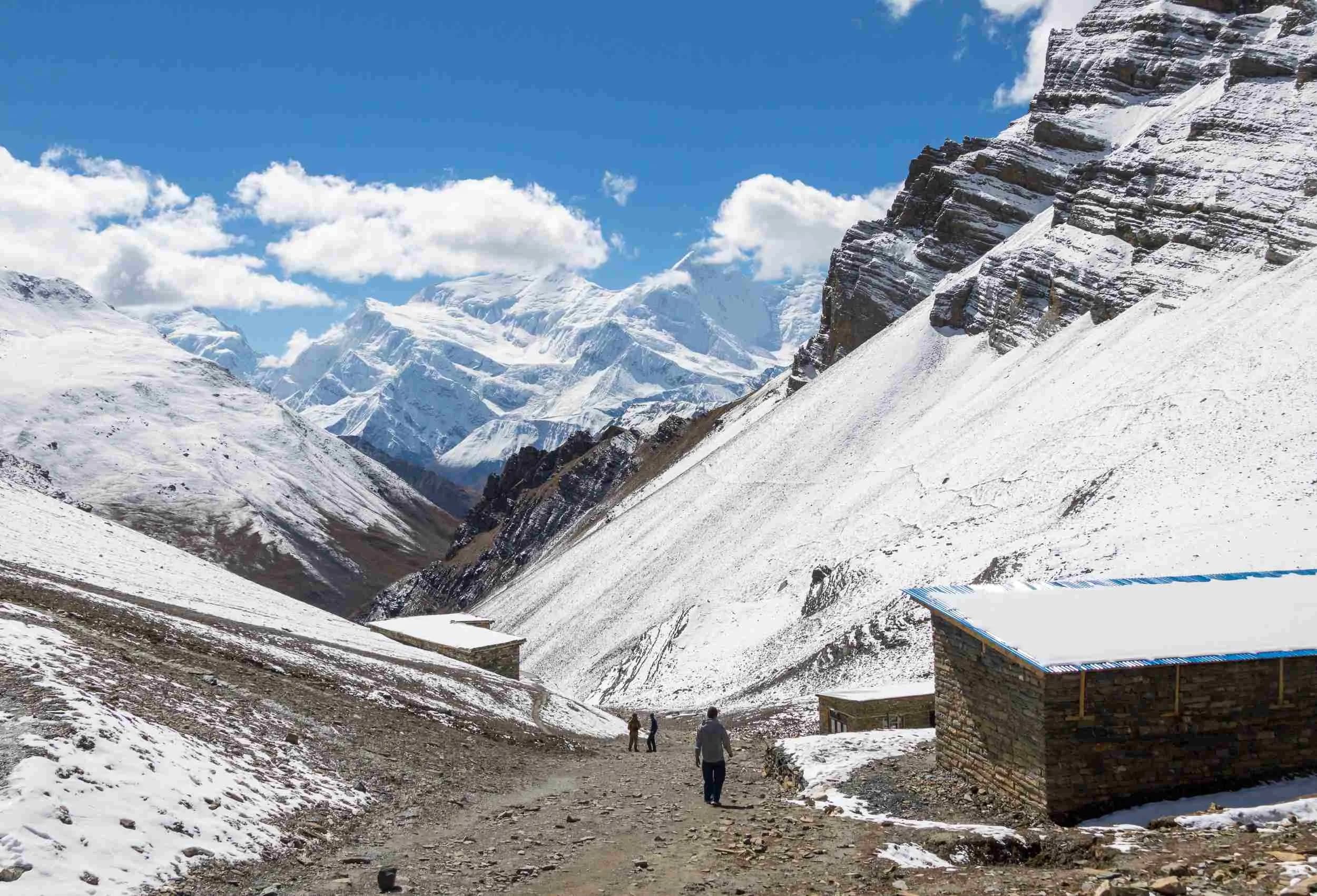 Trekkers walking through a snowy mountain pass with stone buildings and high peaks in the background