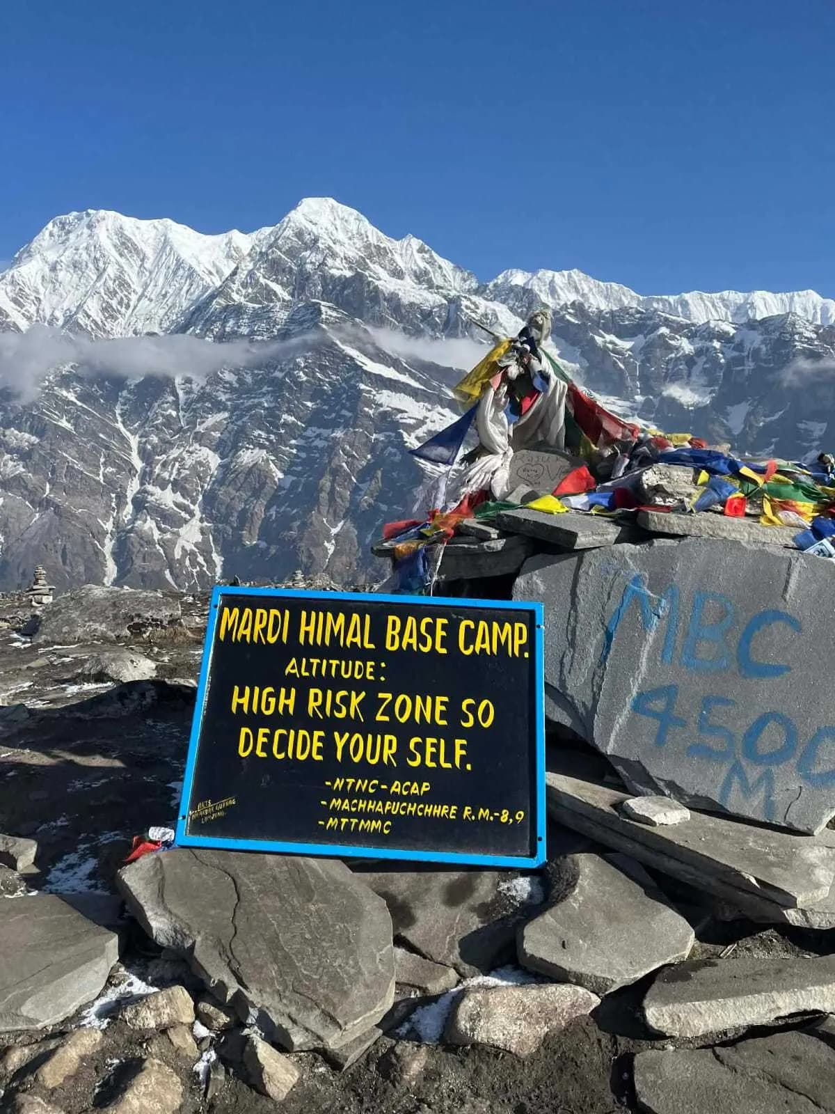 Mardi Himal Base Camp sign with prayer flags and snowy peaks in the background