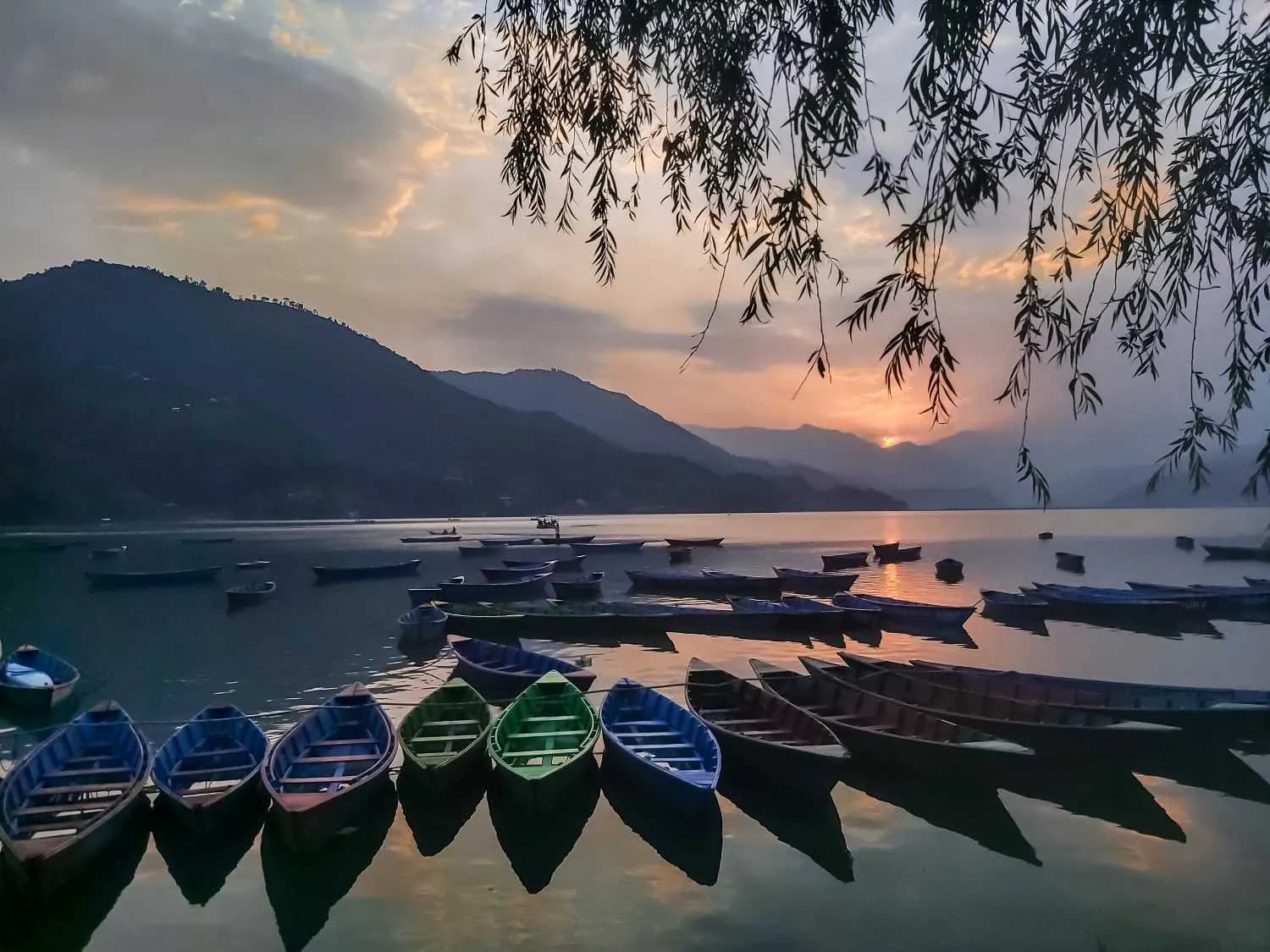 Calm evening at Phewa Lake in Pokhara with colorful boats docked in the foreground and the sun setting behind the silhouettes of the surrounding mountains
