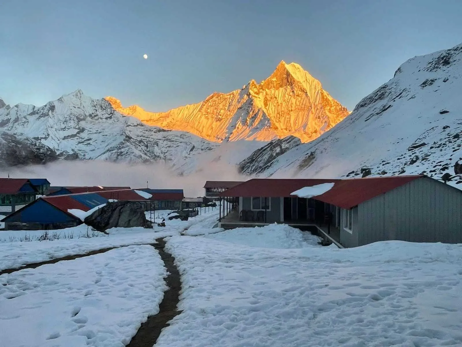 Snow-covered lodges at Annapurna Base Camp with sunrise lighting the Annapurna peaks and a moon visible in the clear Himalayan sky