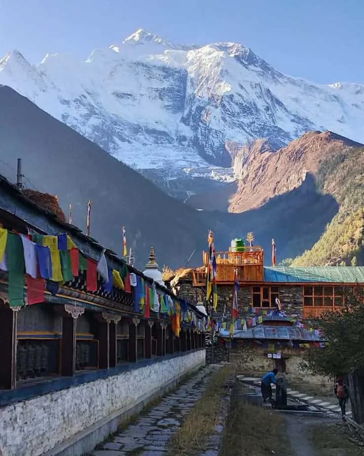 Traditional stone house and prayer flags on a trekking trail with a snowy mountain backdrop in Nepal
