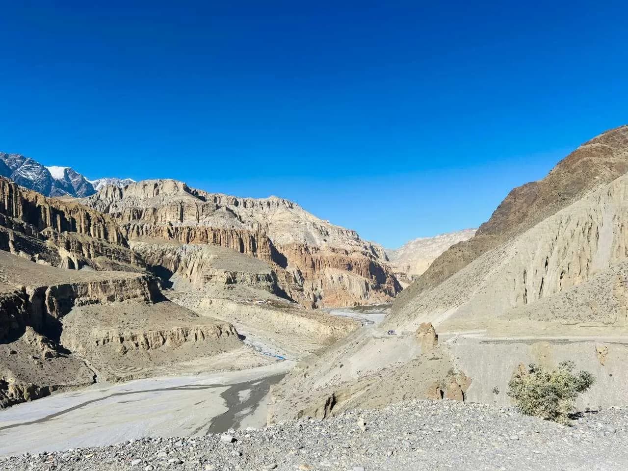 Dramatic eroded cliffs and a wide riverbed winding toward distant snow-capped Himalayan peaks