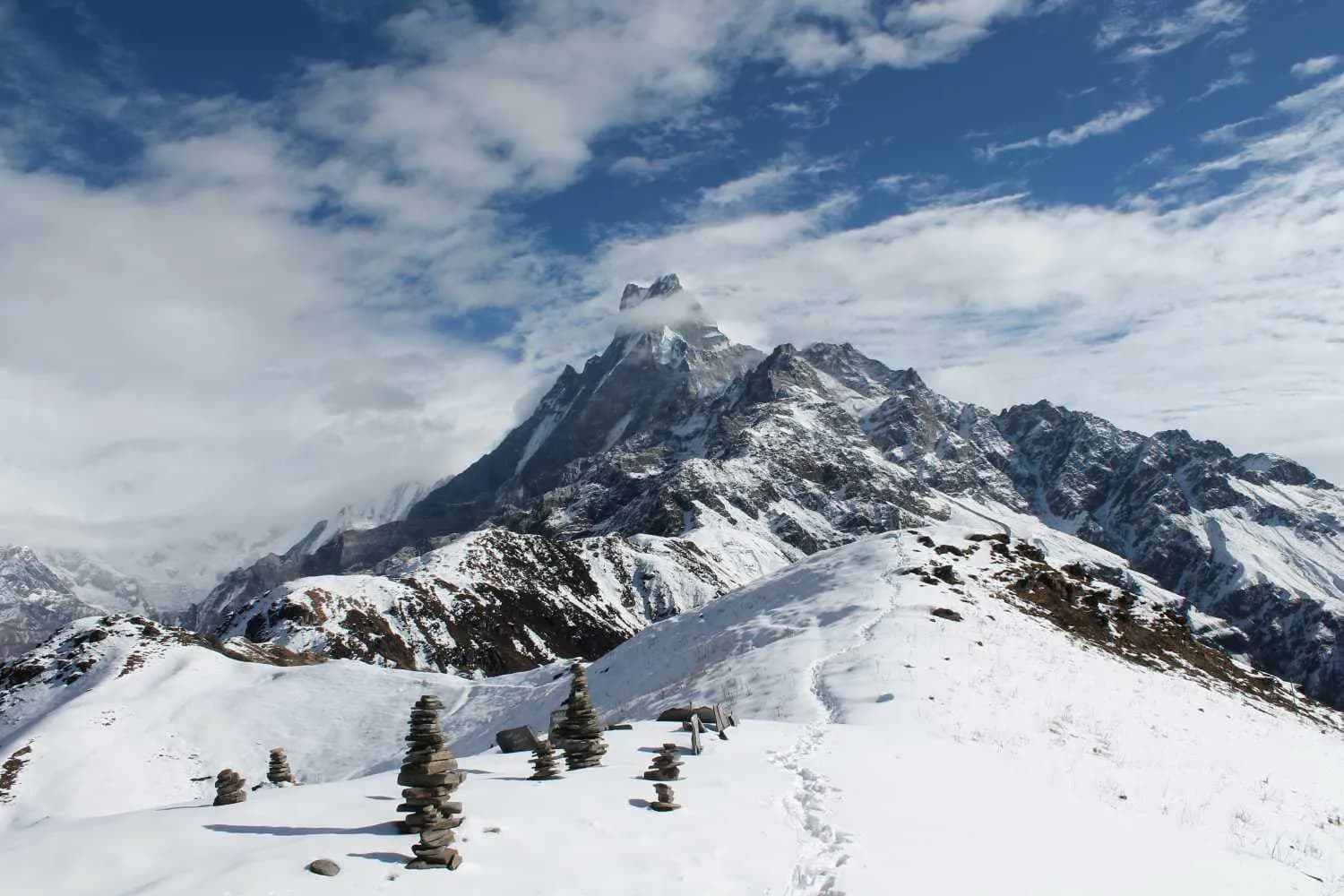 Snowy trekking trail with stone cairns leading toward a cloud-draped Machhapuchhre